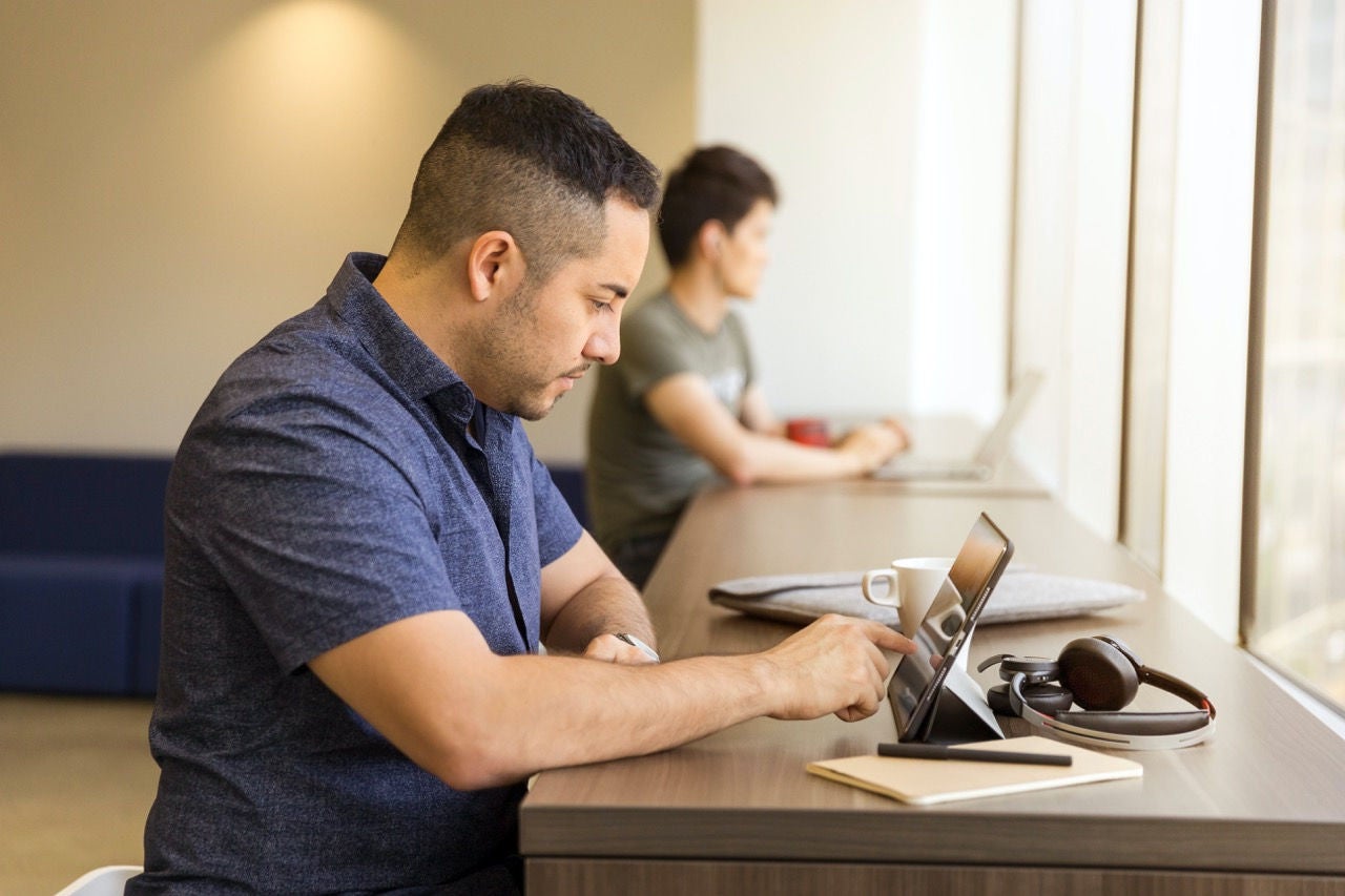 A man in a blue polo shirt sitting at a desk using a tablet with a mug to his left and a notebook and pen to his right.