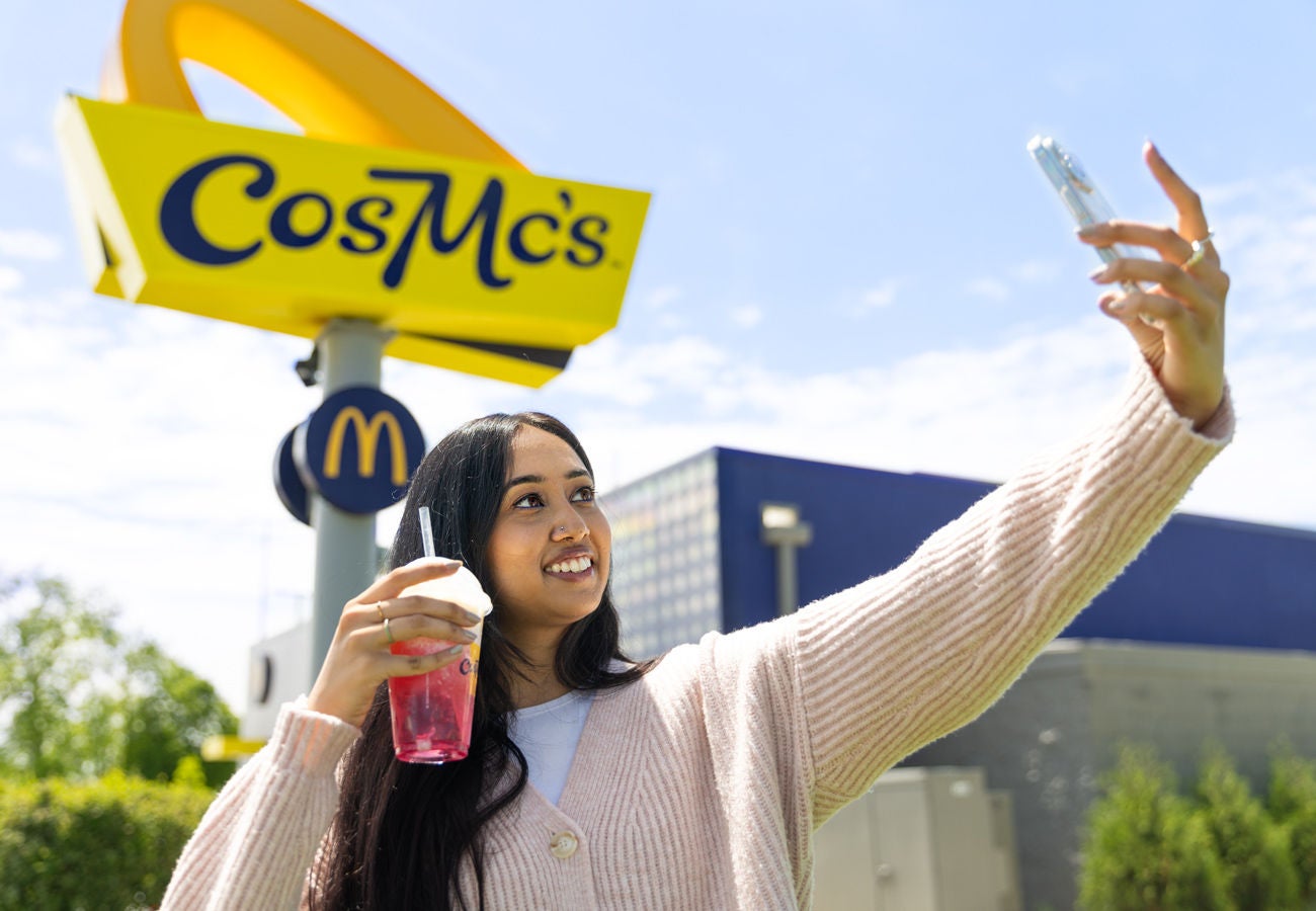 A young woman poses with a CosMc's drink for a selfie in front of the sign on a sunny day.