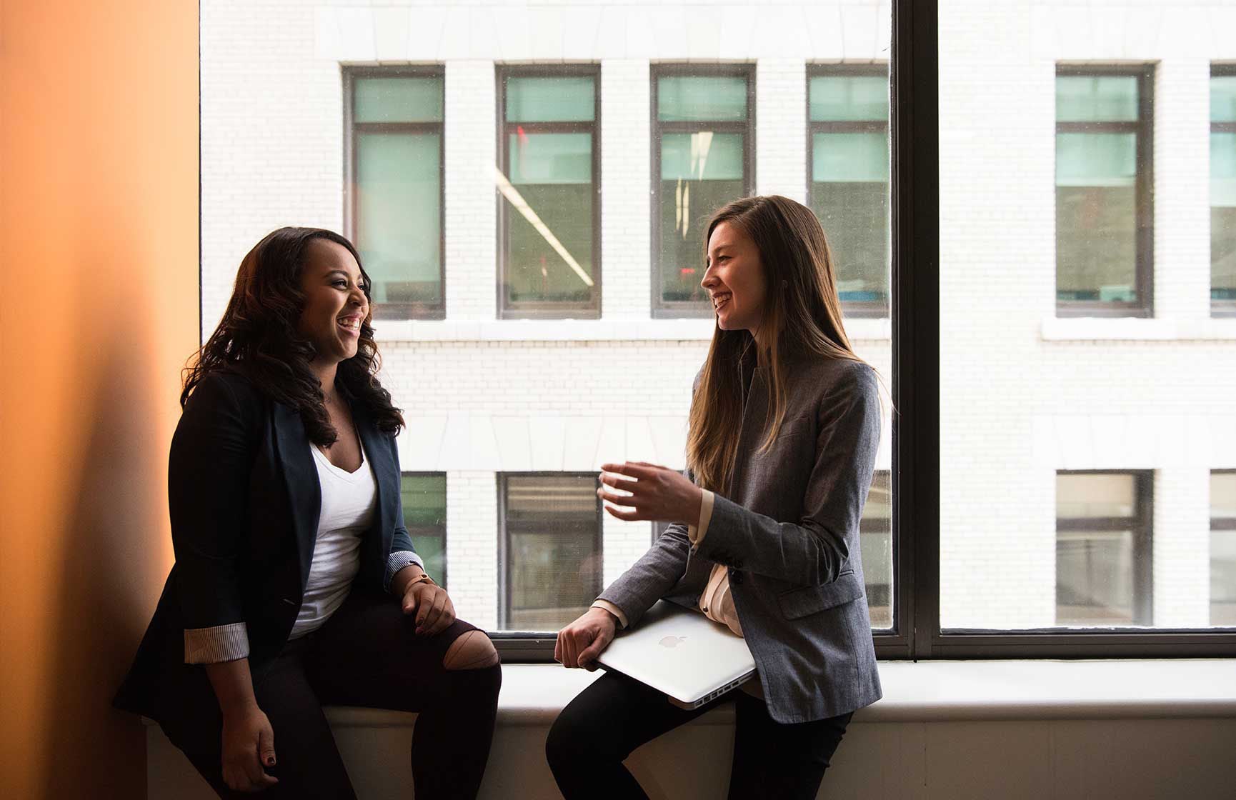 Two smiling women having a conversation sitting by a window.