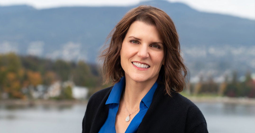 Headshot of Western Canada's general manager, Stephanie Van Eyk, wearing a blue shirt and black jacket, smiling against a blurred, scenic backdrop.