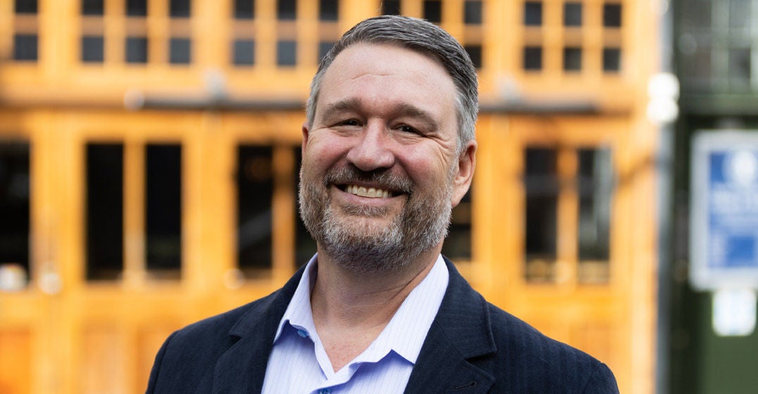 Headshot of London's general manager, Todd Sink, wearing a dark blazer and light blue shirt, smiling with a yellow building in the background.