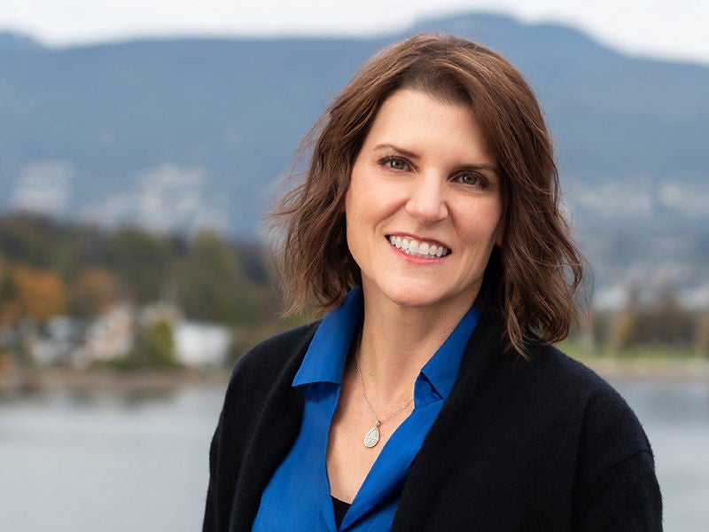 Headshot of Western Canada's general manager, Stephanie Van Eyk, wearing a blue shirt and black jacket, smiling against a blurred, scenic backdrop.