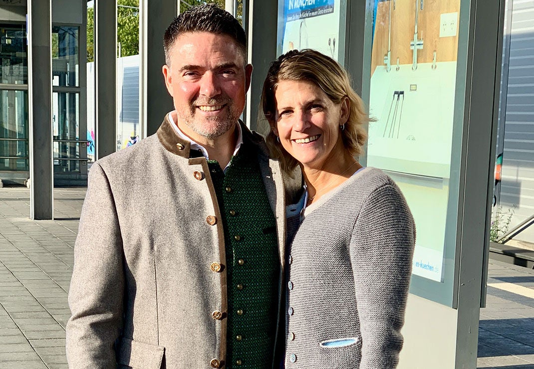 Germany leader, Denis Gassmann, wearing a gray jacket and green patterned vest, standing next to his wife in a gray sweater.