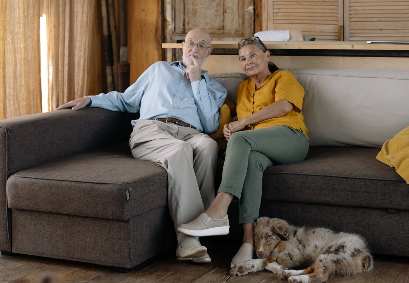 A senior couple and their dog rest on the couch of their home.