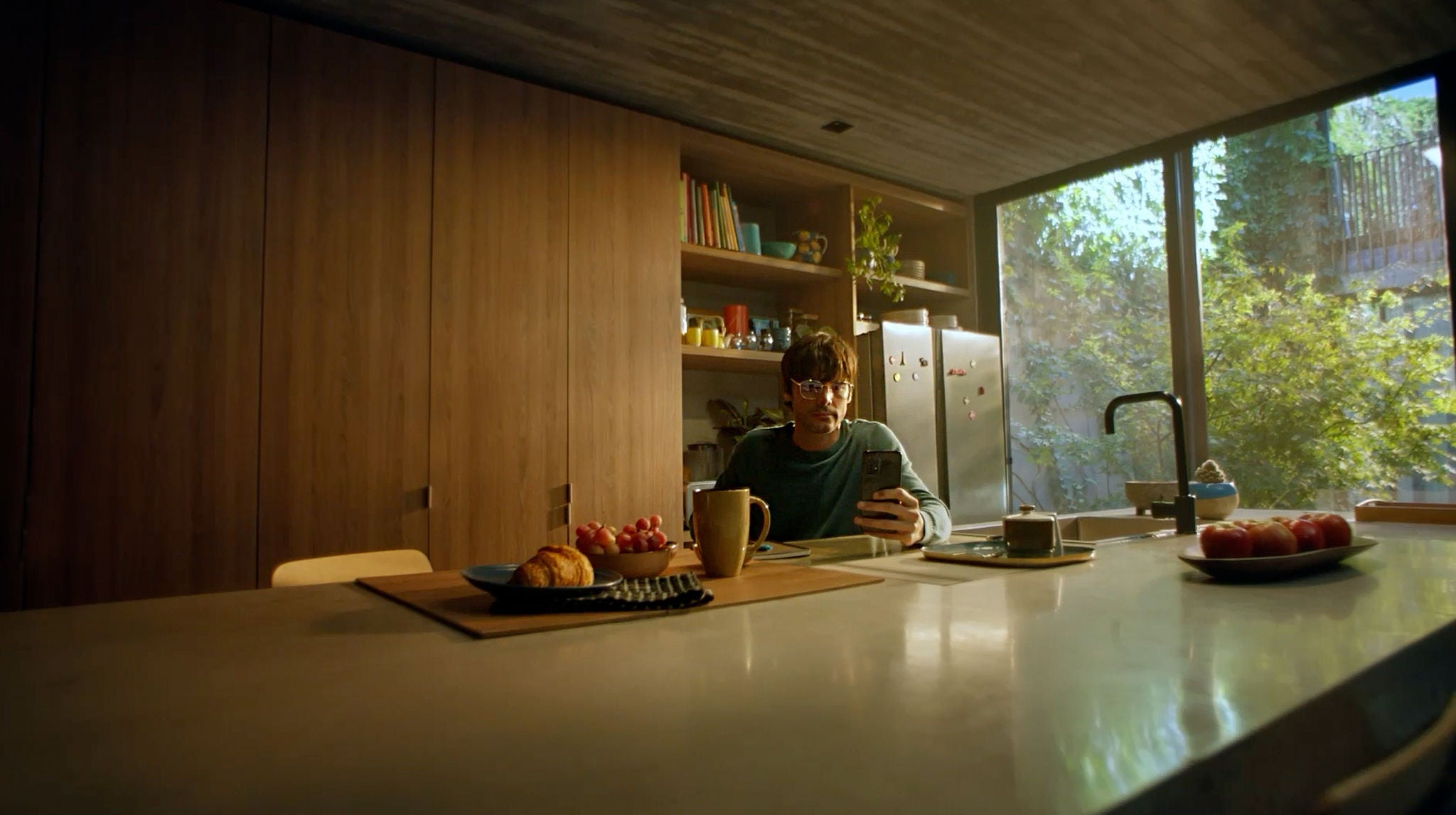 A man sits in a kitchen eating breakfast and looking at his mobile device.