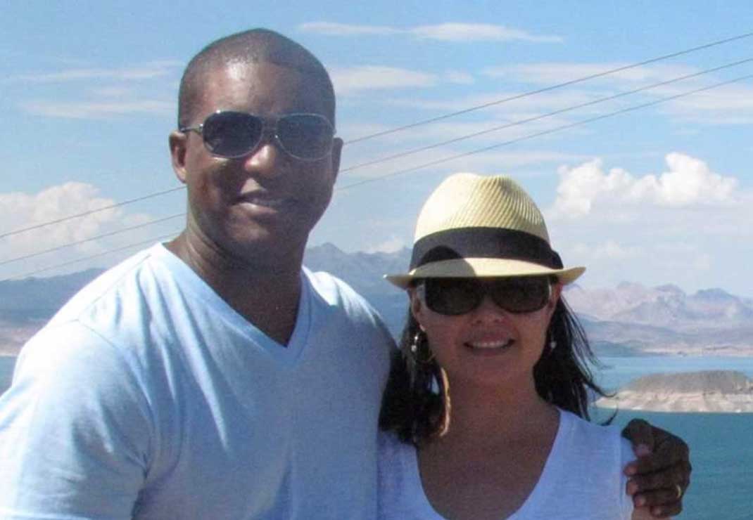 Los Angeles general manager Karl Mehu wearing sunglasses and a white shirt, standing against a scenic ocean backdrop with his wife in a straw hat, sunglasses, and white shirt.